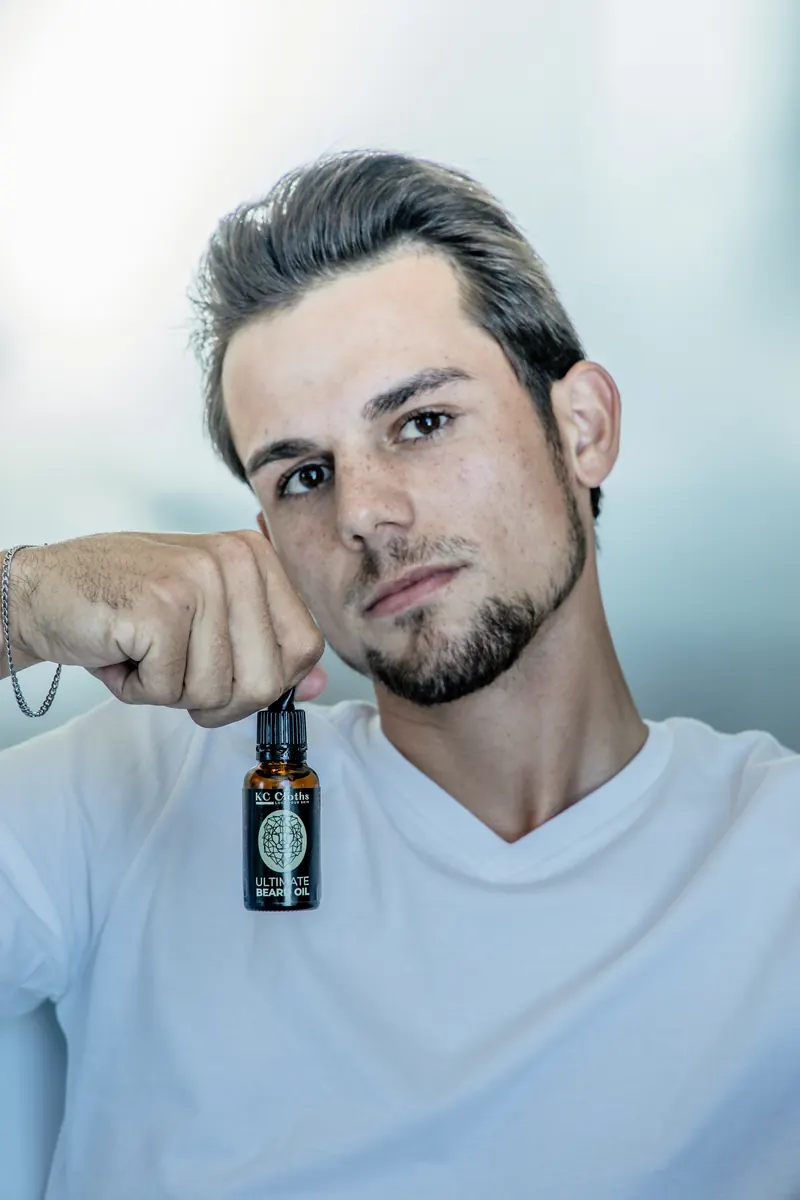 A young man in a white T shirt holding up a bottle of beard oil