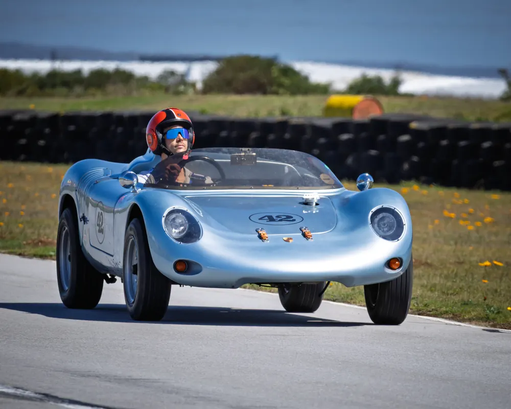 A silver vintage racing car on track, with the driver in a red helmet